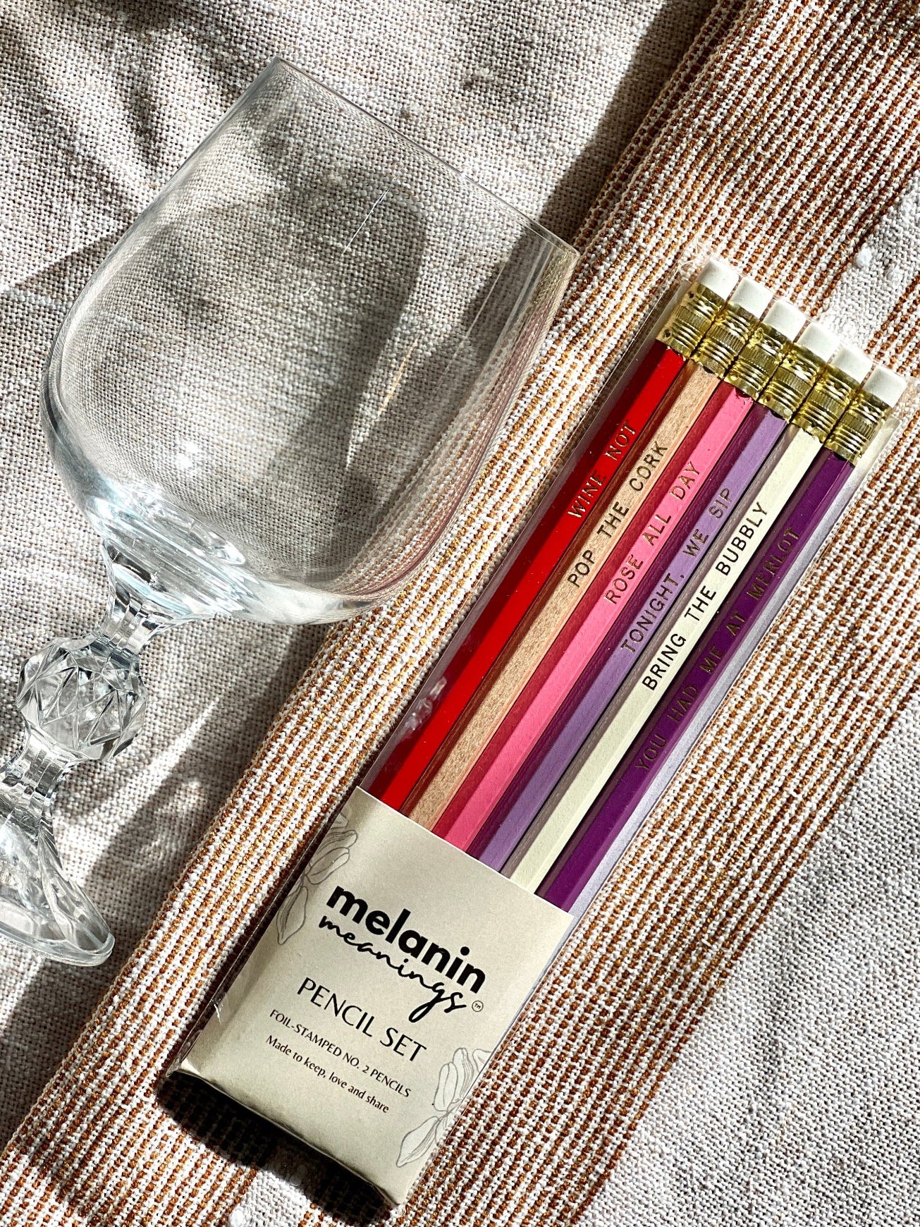 A Melanin Memoirs pencil set with five colored pencils in red, pink, orange, cream, and purple, displayed next to a clear wine glass on a textured beige linen surface.